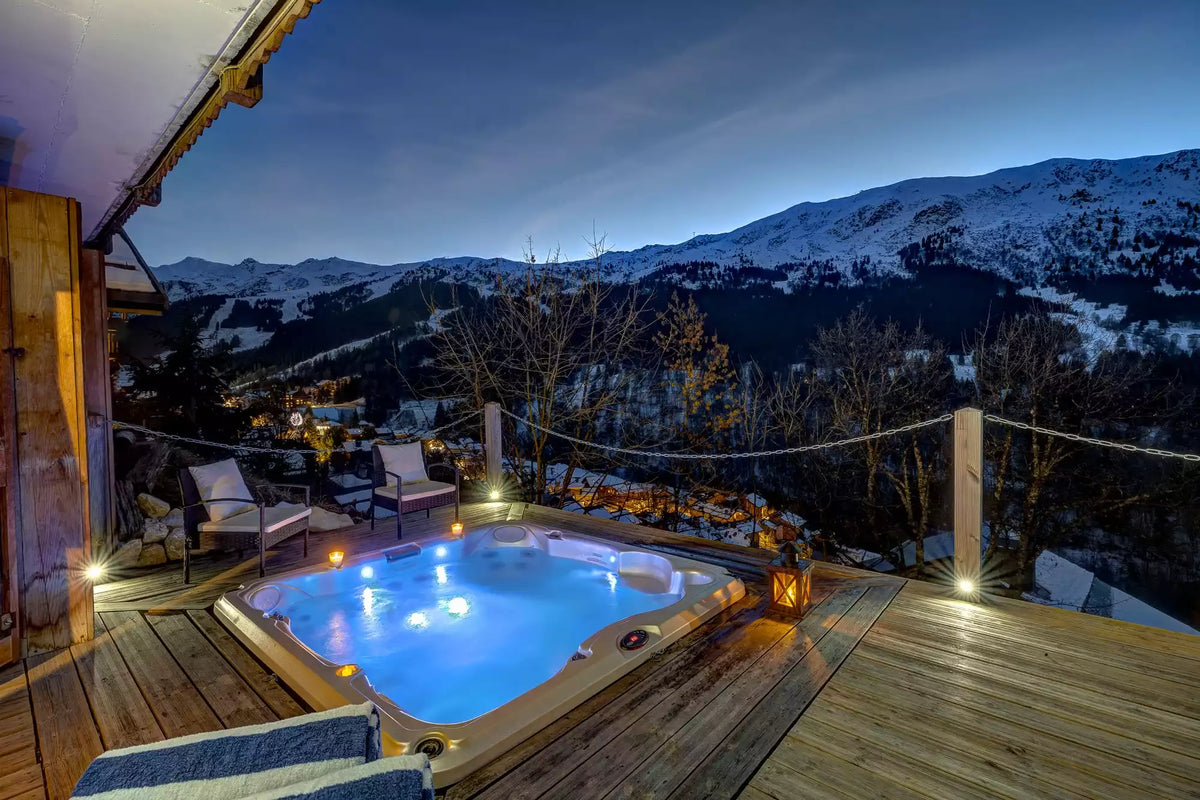A glowing hot tub on a wooden deck at dusk, overlooking a snowy mountain landscape. Two lounge chairs sit nearby, with warm lights and a rope railing separating the deck from the drop-off.