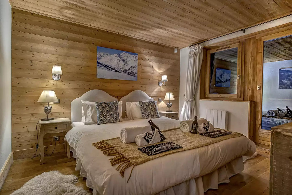A cozy mountain-style bedroom with a large bed dressed in cream bedding, patterned pillows, and a tan throw blanket. The room has light wood paneled walls and ceiling, two bedside tables with lit lamps, a framed mountain landscape print above the bed, and a window looking into an outdoor shower area.