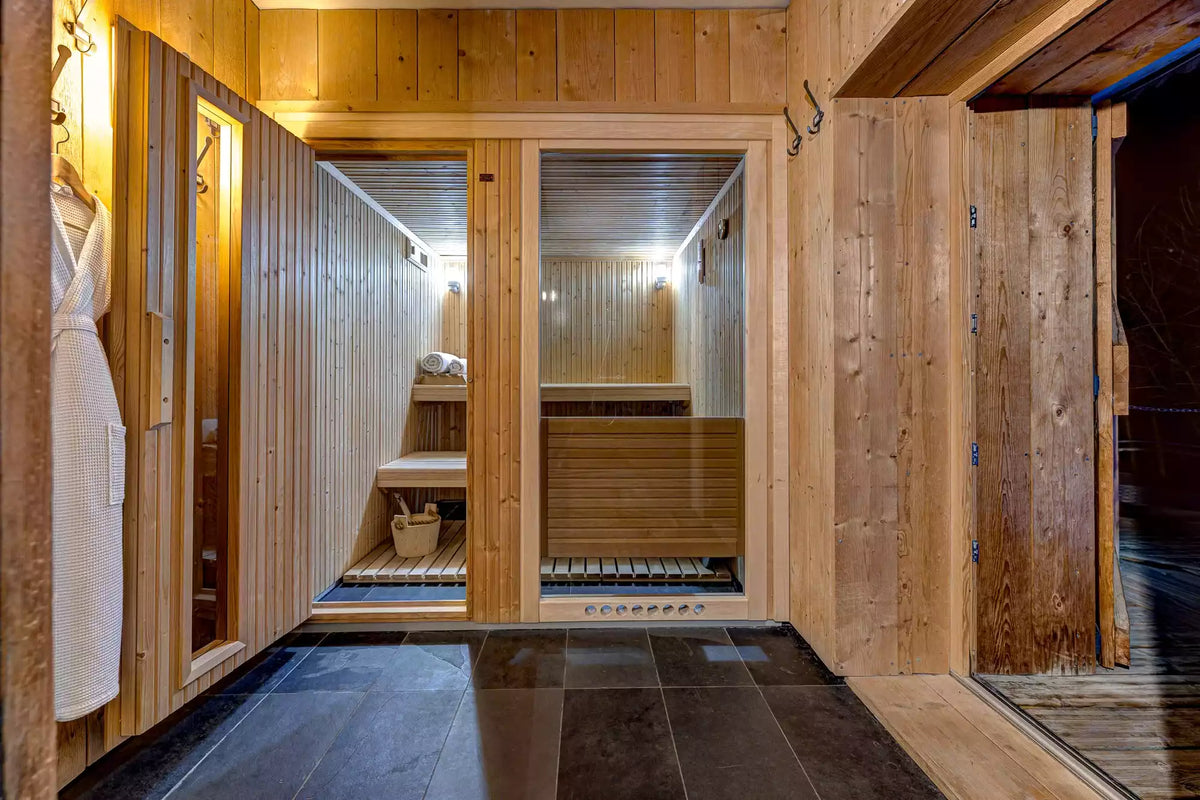 A warm, inviting wooden sauna interior with light wood paneled walls, glass doors, built-in wooden benches, dark stone floor tiles, and a white robe hanging on the left.