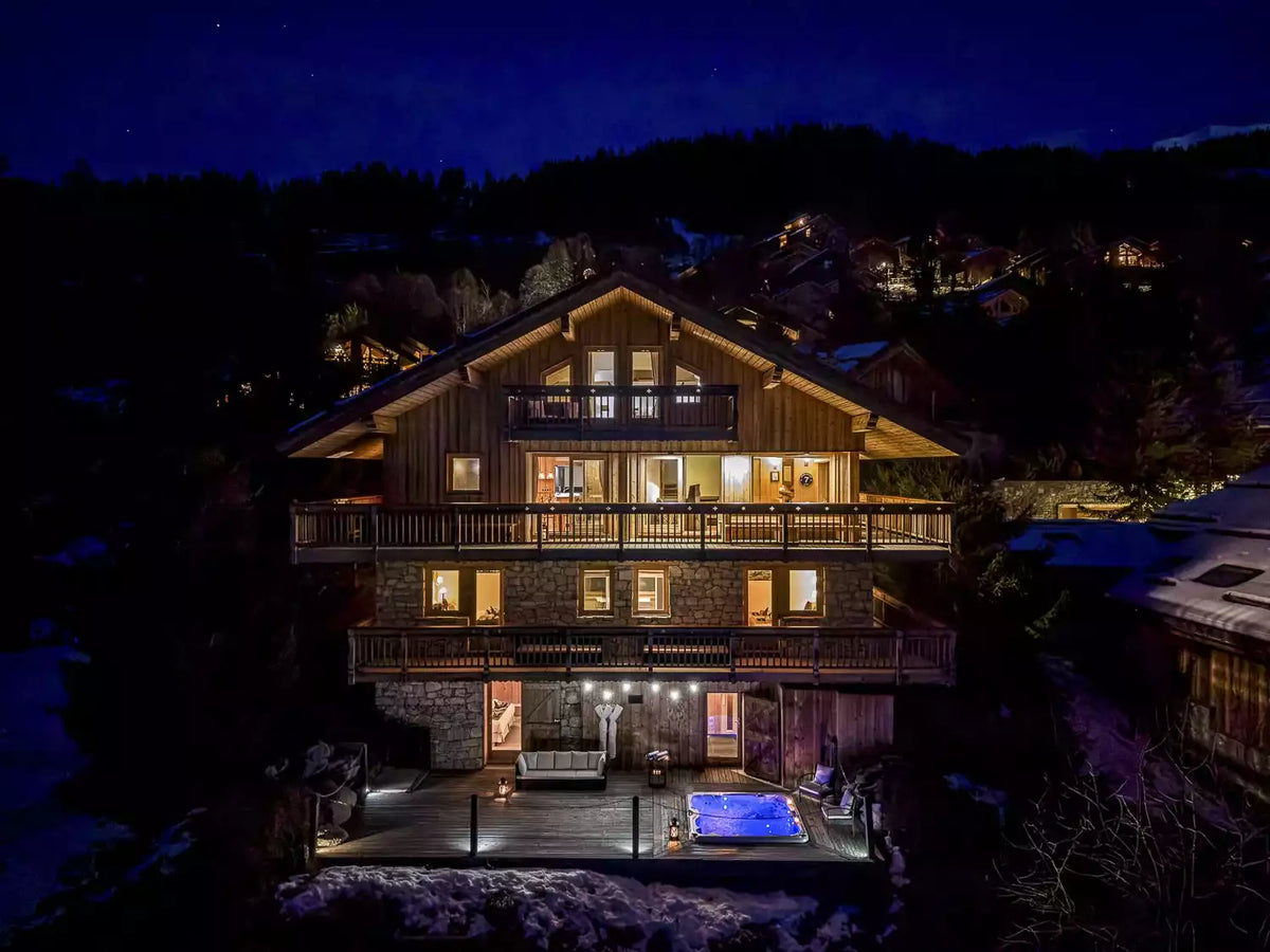 A luxury chalet-style mountain lodge at night, featuring warm golden lights glowing from the windows, a snow-covered patio with an illuminated blue pool, and a dark forested hillside in the background.