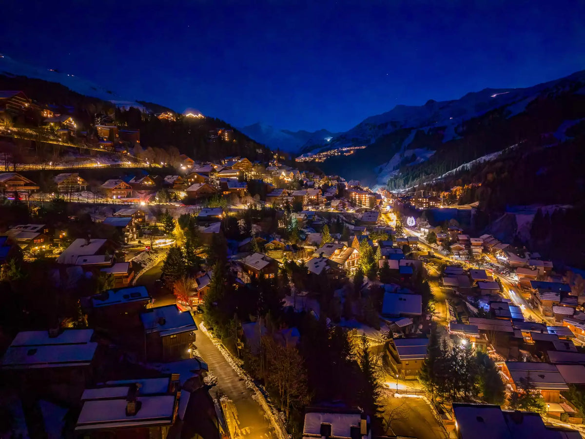 Aerial night view of a snow-covered mountain village with glowing yellow streetlights, clustered buildings, dark trees, and deep blue mountains under a starry sky.