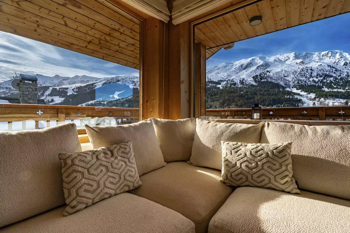 A modern U-shaped sectional sofa in a warm beige fabric with two patterned accent pillows, situated inside a wooden chalet overlooking a snowy mountain landscape.