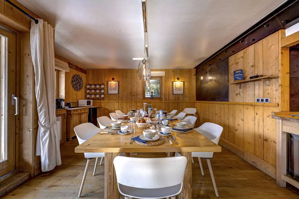 A rustic wooden dining room interior with a long wooden table set for multiple people, surrounded by white chairs. The room features light wood paneled walls, a wooden ceiling, and a hanging light fixture.