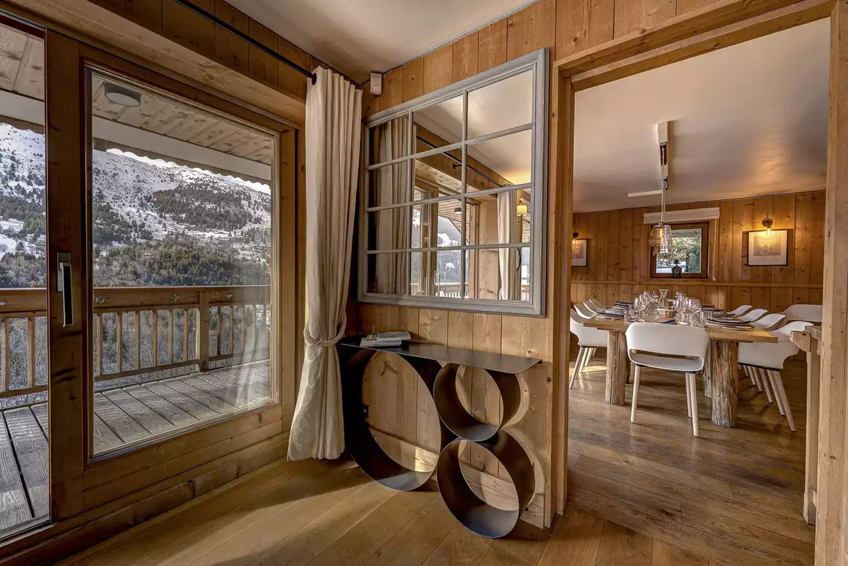 A modern interior entryway with wood-paneled walls, a large wall mirror, and a sculptural black console table. Large glass doors on the left offer a view of snowy mountains, and an open doorway on the right reveals a dining room with a wooden dining table and white chairs.