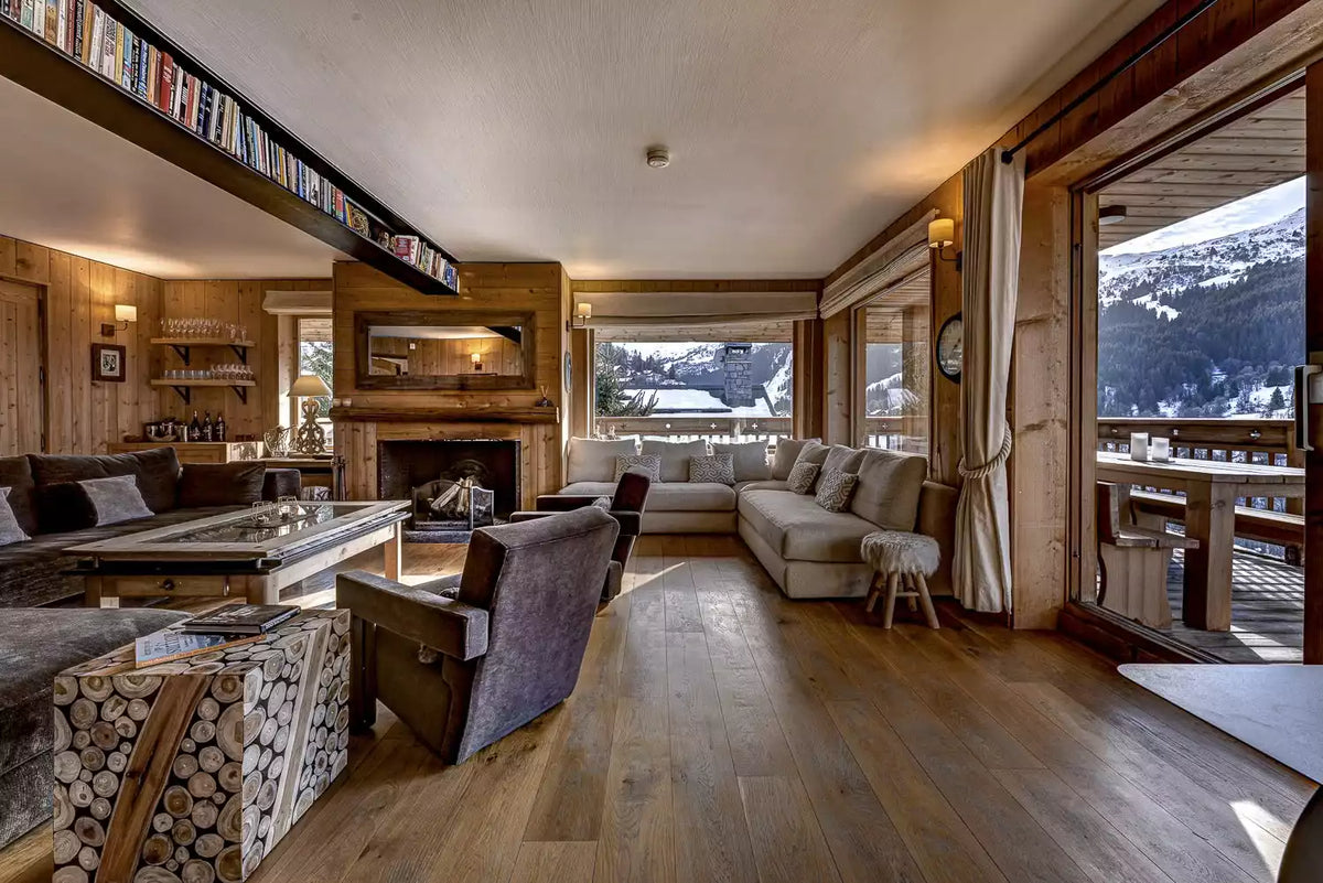 A luxurious ski chalet living room with warm wood paneling and floors, featuring a beige sectional sofa, a dark purple accent chair, a fireplace, and large windows looking out onto a snowy mountain landscape.