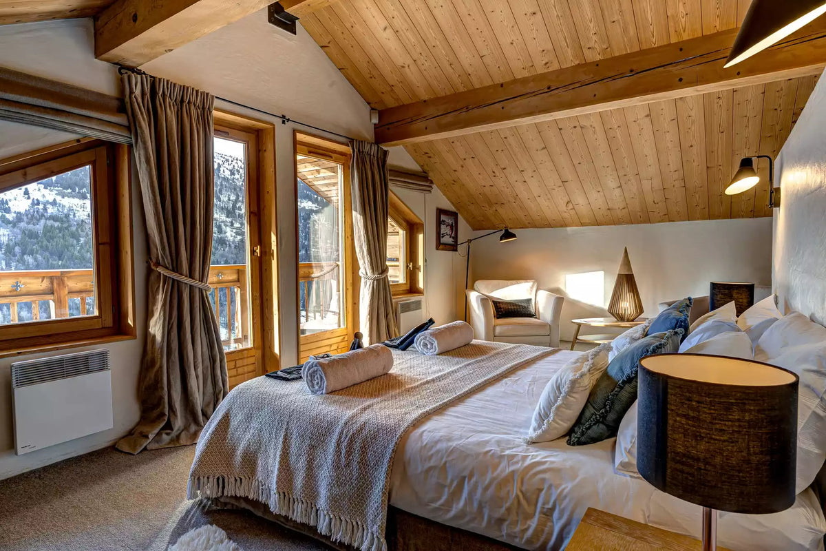 A cozy, rustic mountain chalet bedroom featuring a large bed with white linens and textured beige throws, set under a sloped wooden ceiling with exposed beams. Large windows with brown curtains offer a view of snowy outdoor scenery, and a modern black table lamp sits in the foreground.