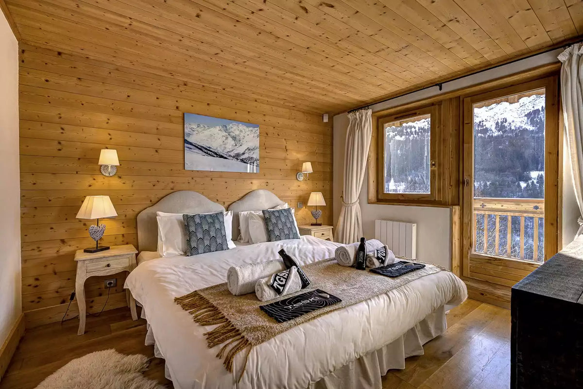 A cozy ski chalet bedroom with a double bed dressed in white linens and blue patterned pillows, set against warm wood-paneled walls. Two bedside tables with lit lamps flank the bed, and large windows with white curtains look out onto a snowy mountain view.