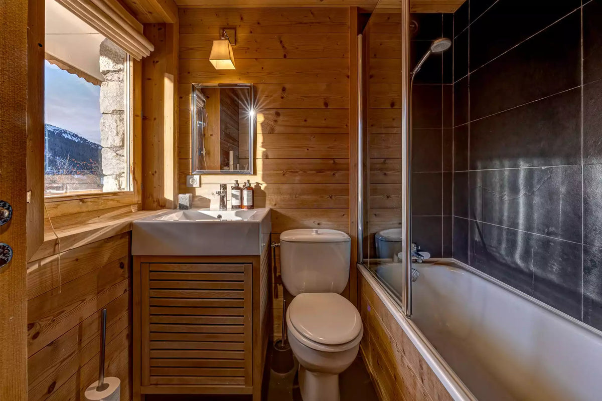 A compact mountain-style bathroom with wood-paneled walls, a white sink vanity, toilet, bathtub with dark tiled surround, and a window showing a snowy mountain view.