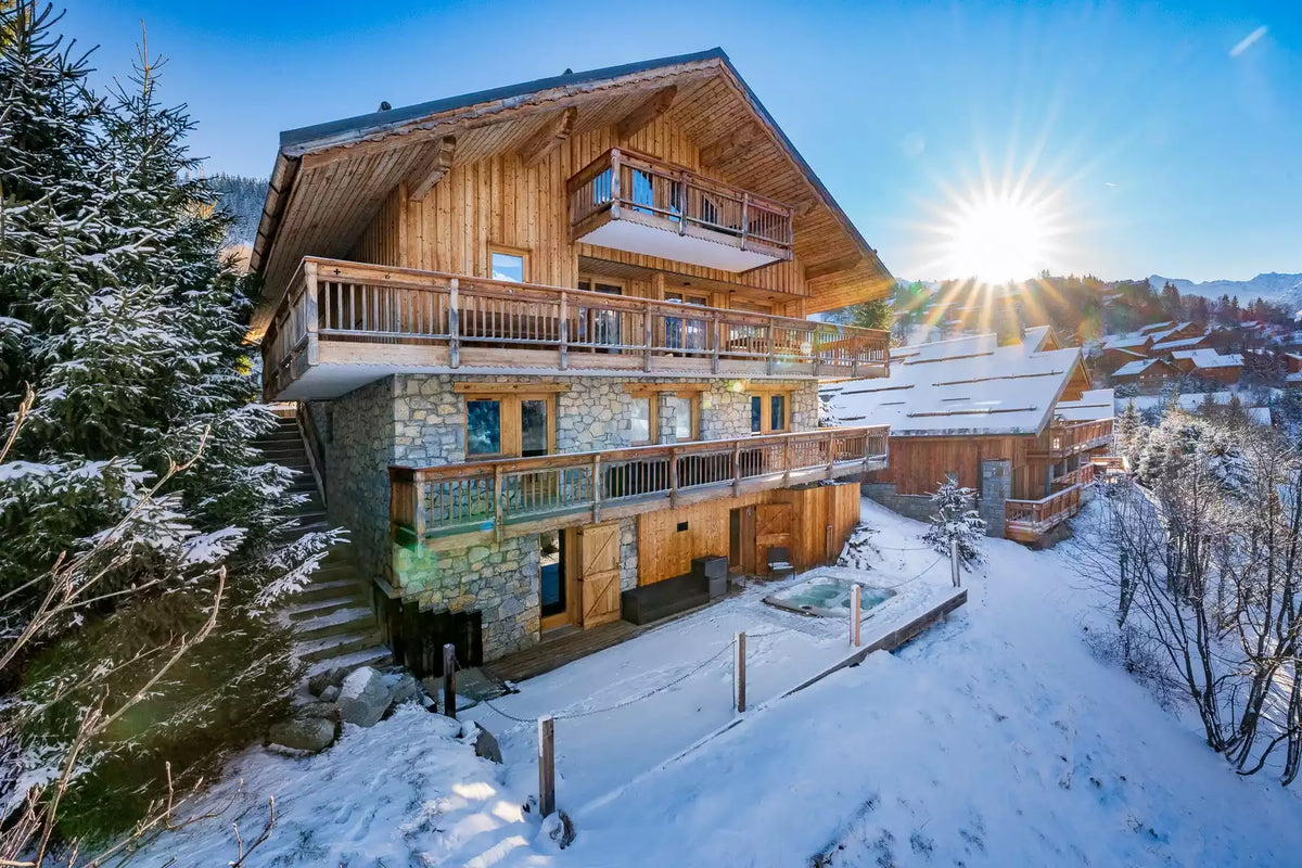 A large alpine chalet with wooden balconies and a stone lower facade, surrounded by snow-covered trees and mountains under a bright sun in a blue winter sky.