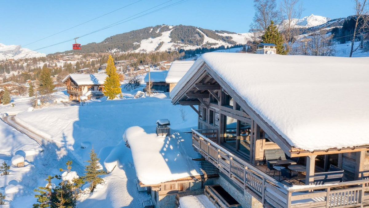 A snowy mountain chalet with a large snow-covered roof and wooden balcony, set in a ski resort with mountains, trees, and a ski lift in the background.