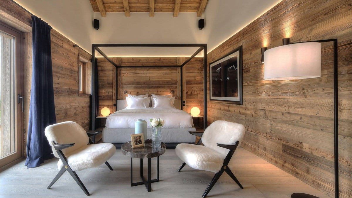 A luxury bedroom interior with a four-poster bed, wood-paneled walls, two white armchairs, a small round coffee table, and a large floor lamp with a white shade.