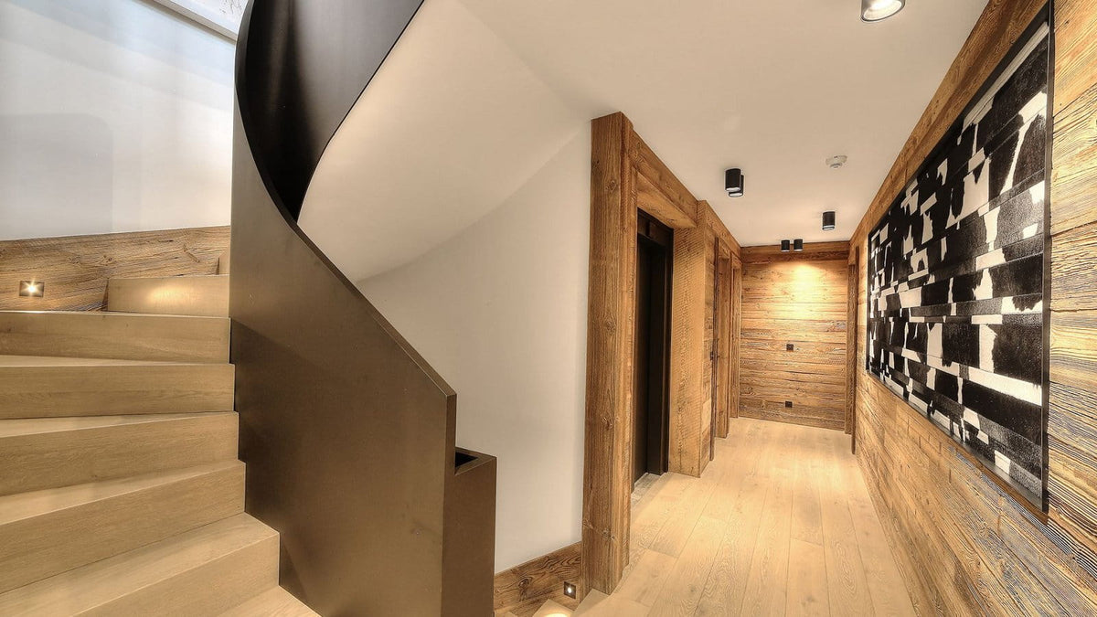 A modern interior hallway with light wood flooring, wood-paneled walls, a spiral staircase on the left, and a large black-and-white abstract wall art piece on the right.