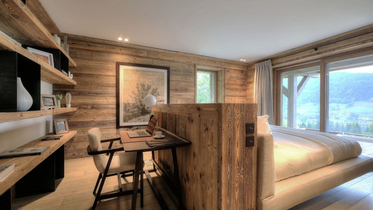 A modern mountain chalet bedroom featuring a low platform bed with cream bedding, a built-in wooden desk and shelving unit with a chair, and a large framed landscape print on a wood-paneled wall. Large windows offer a scenic view of the alpine valley.