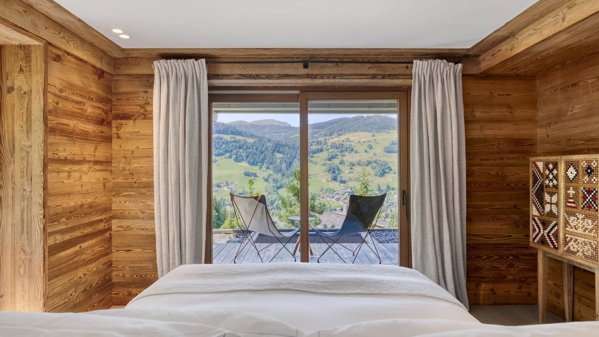 A rustic wooden bedroom with a large bed in the foreground, sheer curtains, and a sliding glass door overlooking a mountain landscape. Two modern chairs sit on the balcony.
