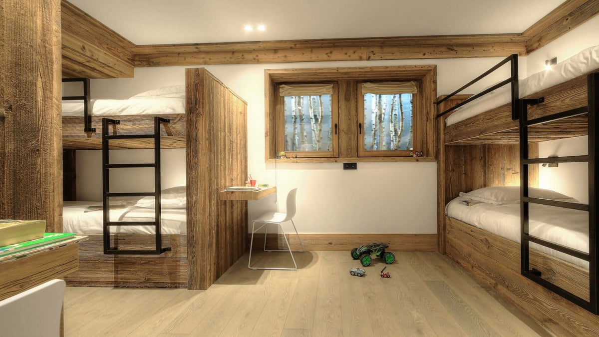 A modern rustic bedroom featuring multiple wooden bunk beds with black metal frames, a small white desk and chair, and a window looking out onto a forest.
