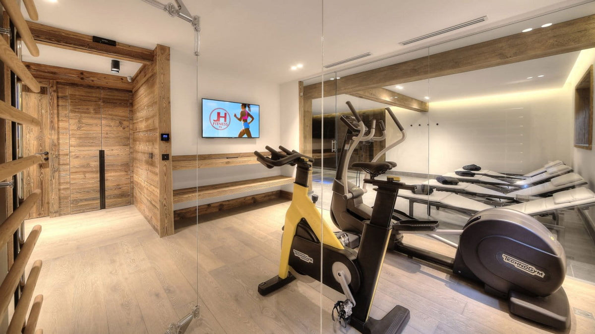 A modern indoor gym space with wooden walls and flooring, featuring a yellow and black stationary bike in the foreground, an elliptical machine, treadmills, and a wall-mounted TV.