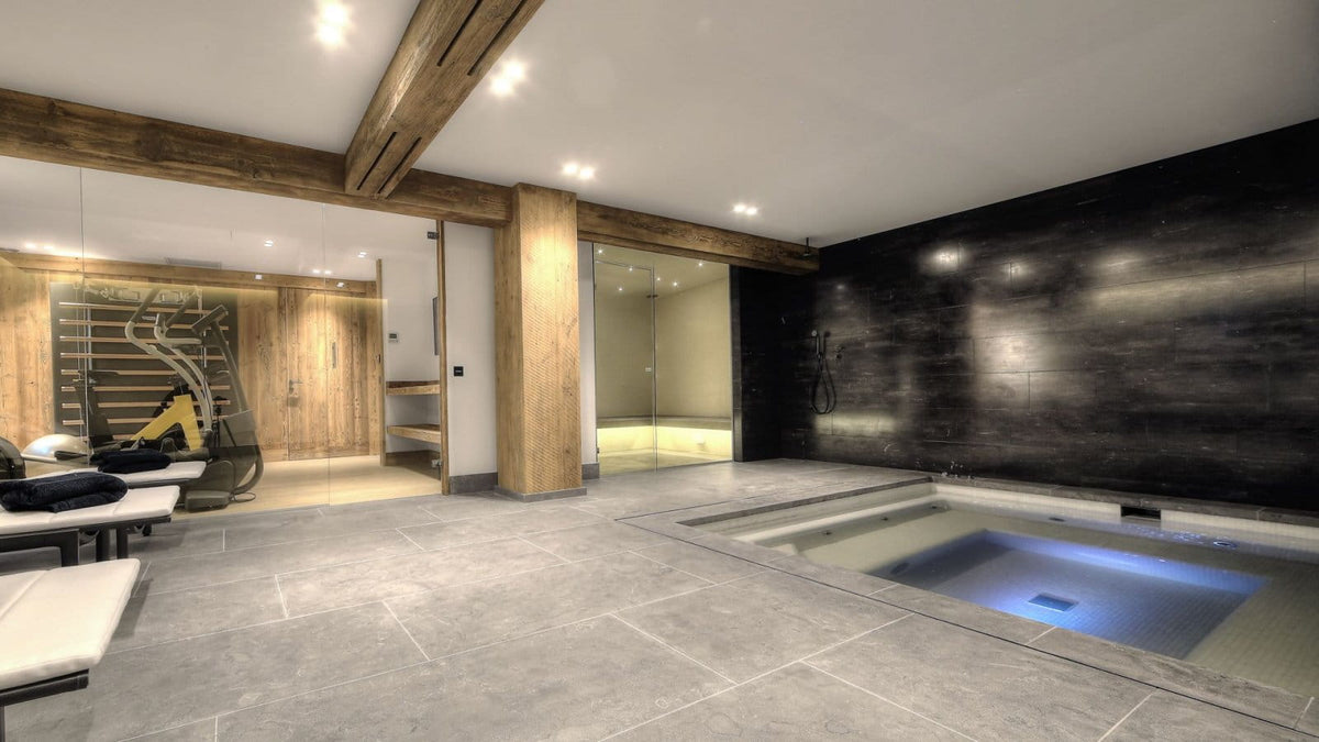 A luxury indoor spa and wellness center featuring a rectangular hot spring or jacuzzi pool with blue water, a large stone-tile floor, a dark wood-paneled wall, and rustic wooden ceiling beams.
