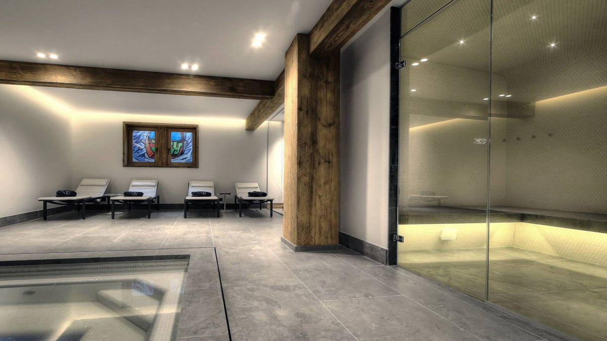 A luxury spa interior with a glass-walled sauna, gray stone floors, wooden beams, and three lounge chairs with towels in a relaxation room.