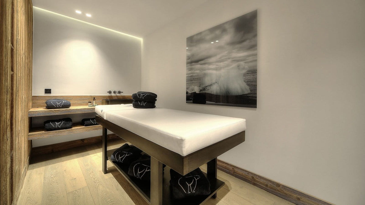 A spa treatment table with a white cushion and dark wood frame, set in a minimalist wellness room with light wood flooring, a large black-and-white wall art print, and neatly folded black towels.