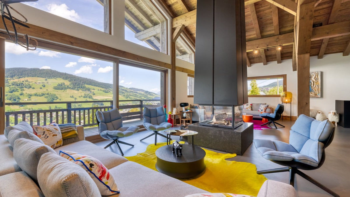 A luxury modern mountain chalet living room with a large beige sectional sofa, blue lounge chairs, a yellow rug, a black fireplace, and a massive wood-beamed ceiling with scenic mountain views outside.