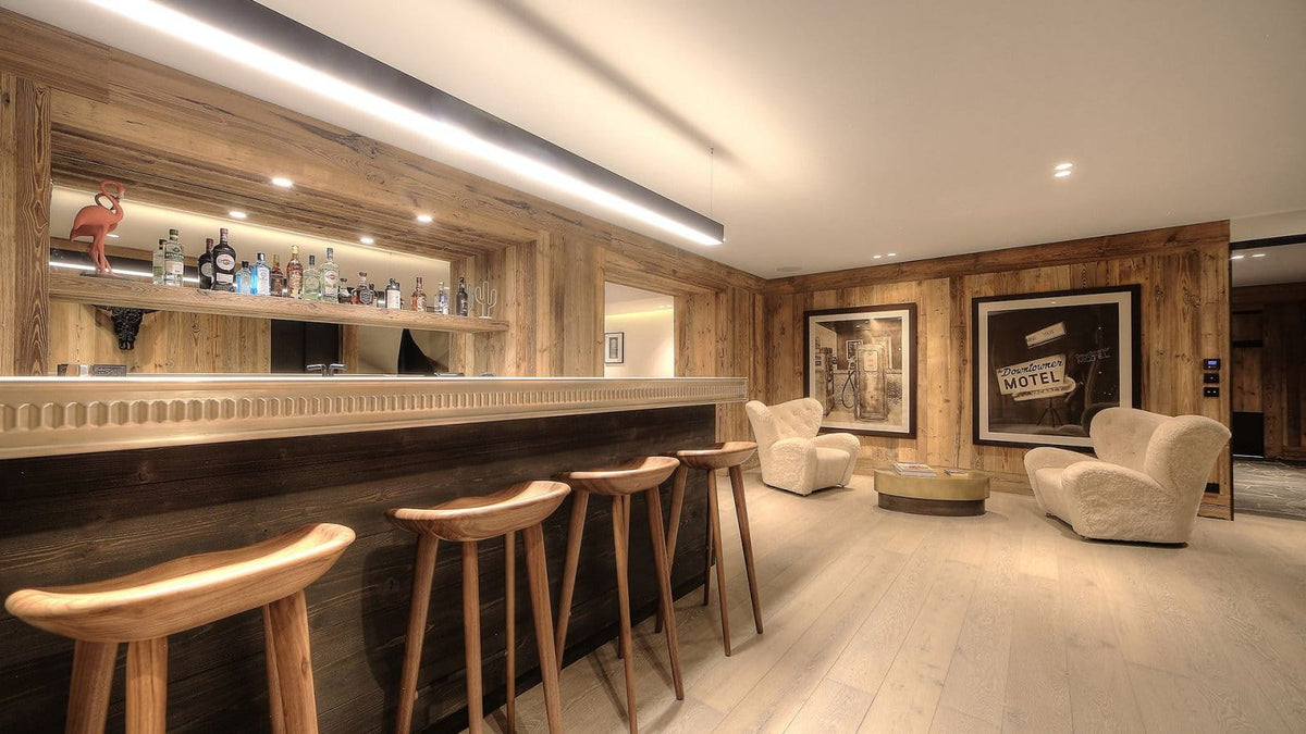 A modern bar and lounge area with a long wooden counter, four wooden bar stools, built-in shelving with bottles, cream armchairs, framed wall art, and light wood flooring.