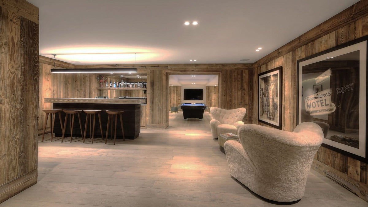 A modern interior lounge or bar area with wood-paneled walls, a bar counter with stools, and two plush cream boucle armchairs in the foreground. Framed wall art hangs on the right, and a doorway leads to another room in the background.
