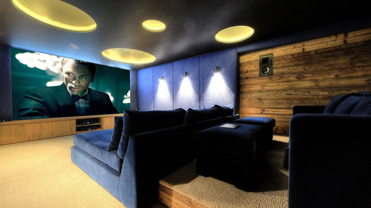 A home theater room with dark blue velvet sectional movie theater seating, a large screen displaying a man in a suit, a wood-paneled wall, and circular ceiling lights.