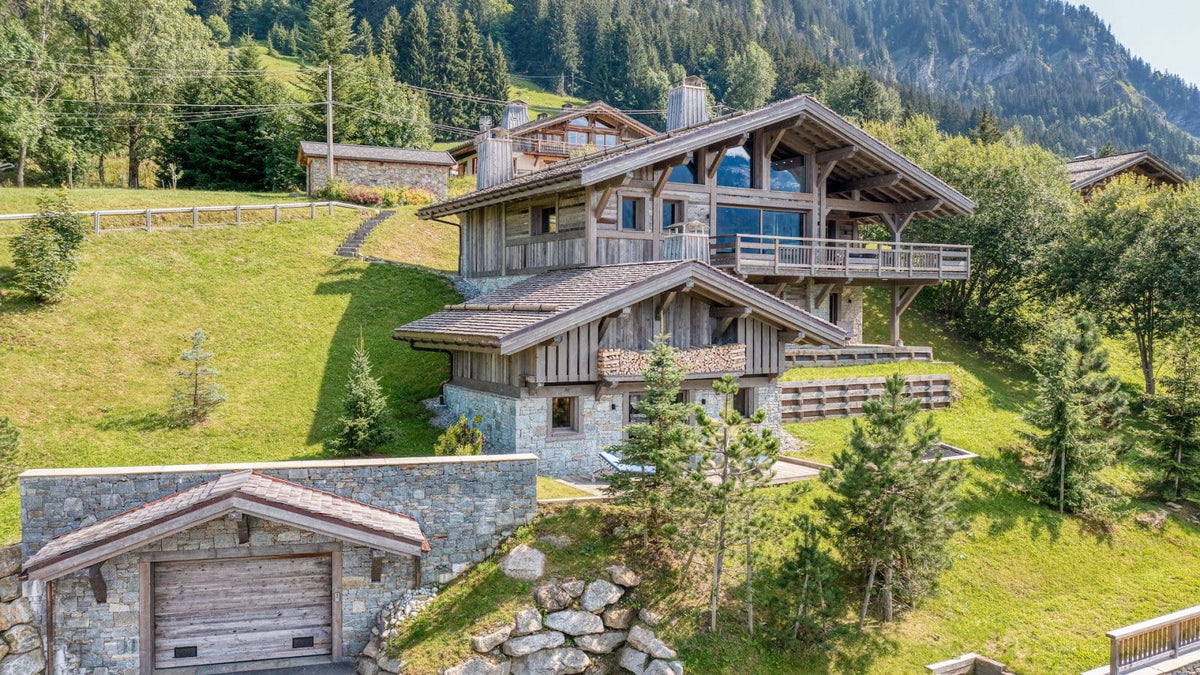 A large alpine-style wooden chalet with stone foundations and a detached stone garage, situated on a grassy hillside with dense evergreen trees and mountains in the background.