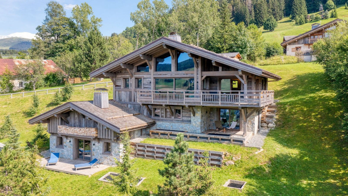 A large modern alpine chalet with wooden and stone exterior walls, large glass windows, and a wraparound balcony, situated on a grassy hillside surrounded by trees.