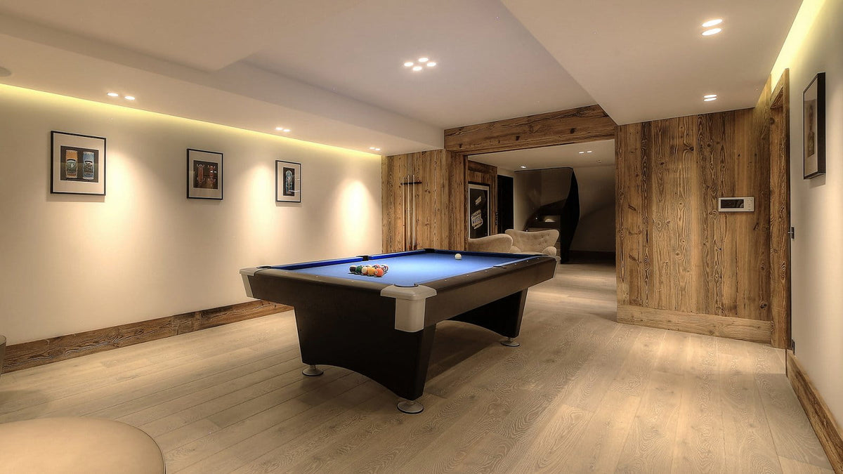 A modern game room featuring a blue pool table in the center, set against light wood-paneled walls and a light wood floor. The room is illuminated by recessed ceiling lights and warm under-cabinet lighting.
