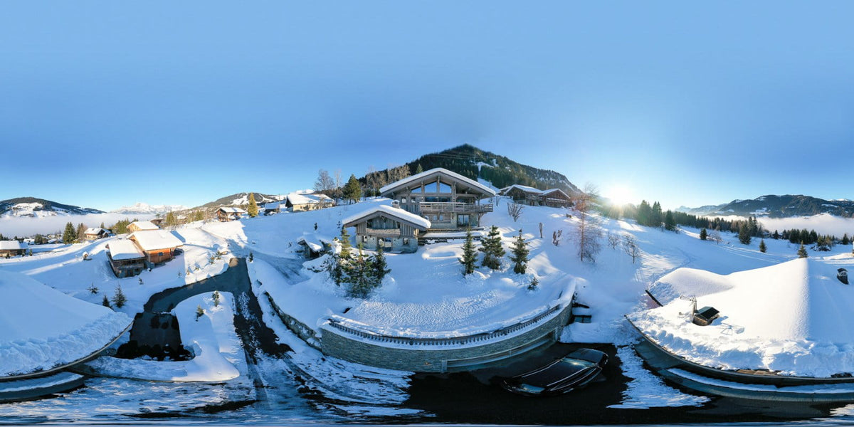 A panoramic winter mountain landscape with snow-covered chalets, evergreen trees, and a bright sun breaking through the blue sky.