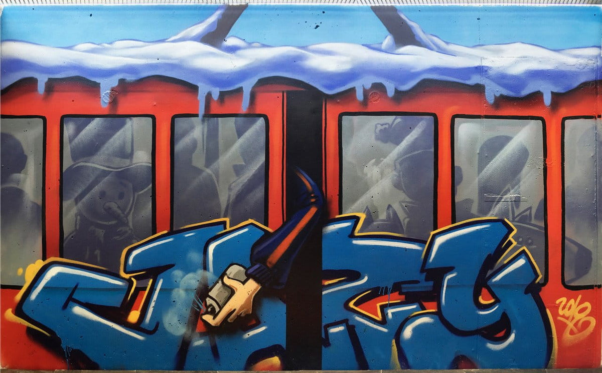 A colorful graffiti mural painted on a red train car, featuring large blue and yellow stylized letters across the front, snow dripping over the top, and gray figures visible through the windows.