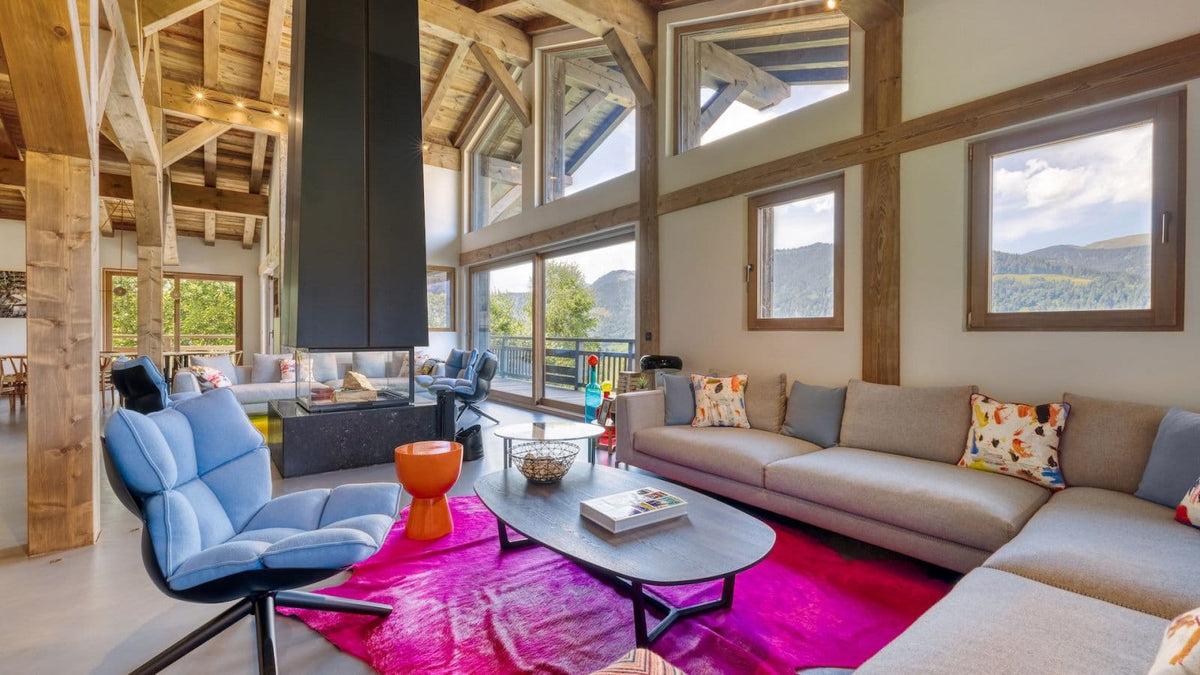 A modern high-ceilinged living room with exposed wood beams, large windows, and a mountain view. The space includes a beige sectional sofa with patterned pillows, a blue lounge chair, a black fireplace, a dark coffee table, and a vivid pink rug.