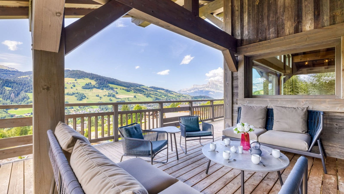 Alpine-style outdoor patio furniture set on a wooden deck with a scenic mountain valley view. The set includes a large beige cushioned sofa, two blue armchairs, a round gray coffee table with white cups and a small pink vase, and the background shows green hills and blue sky.
