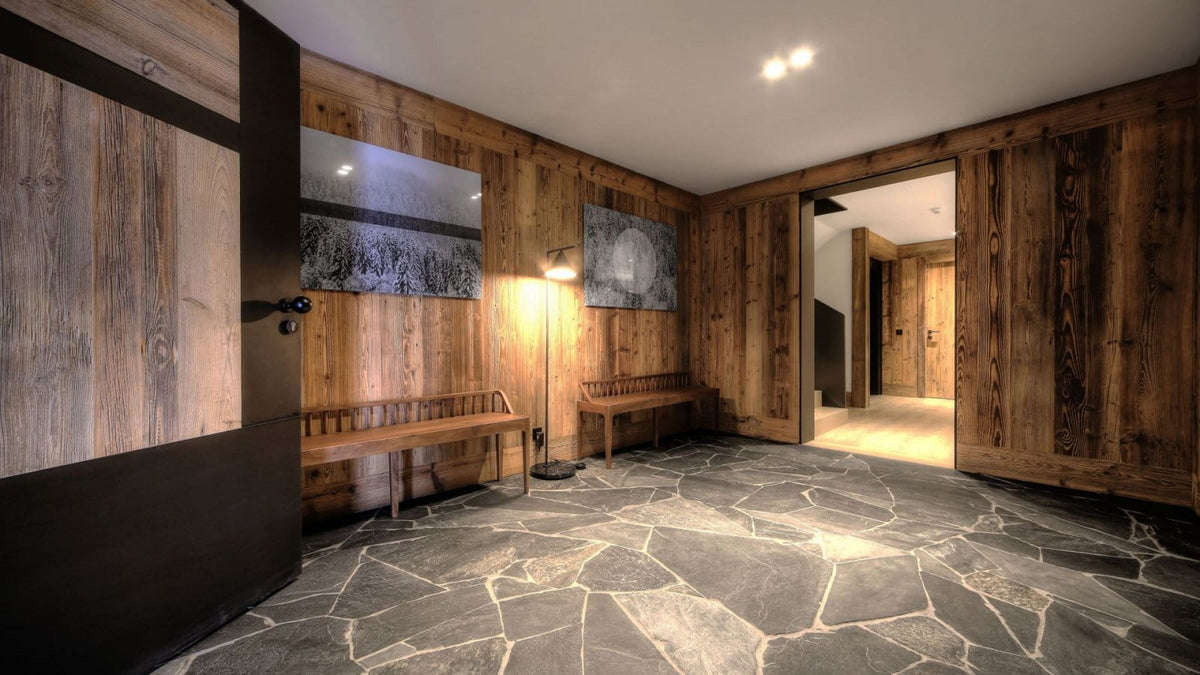 A rustic interior space with wood-paneled walls, a stone-tiled floor, wooden benches, wall art, and a floor lamp, shown as an architectural or cabin interior scene.