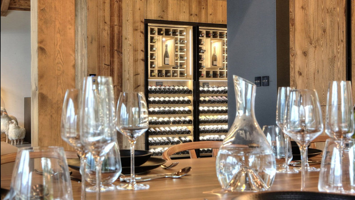 A dining table set with multiple clear wine glasses and a glass decanter in the foreground, with a wooden wine cabinet filled with bottles in the background against a rustic wood-paneled wall.