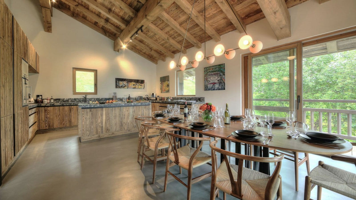 A rustic modern kitchen and dining room interior with exposed wooden ceiling beams, a long dining table set with black plates and glassware, wooden chairs, a kitchen island, and large windows overlooking green trees.