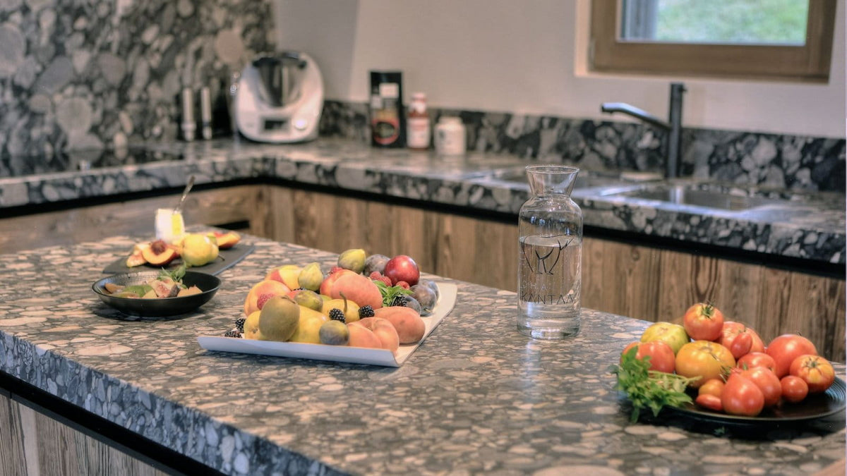 A modern kitchen scene with a granite countertop displaying a fruit platter, a bowl of tomatoes, a glass carafe of water, and other kitchen items in the background.