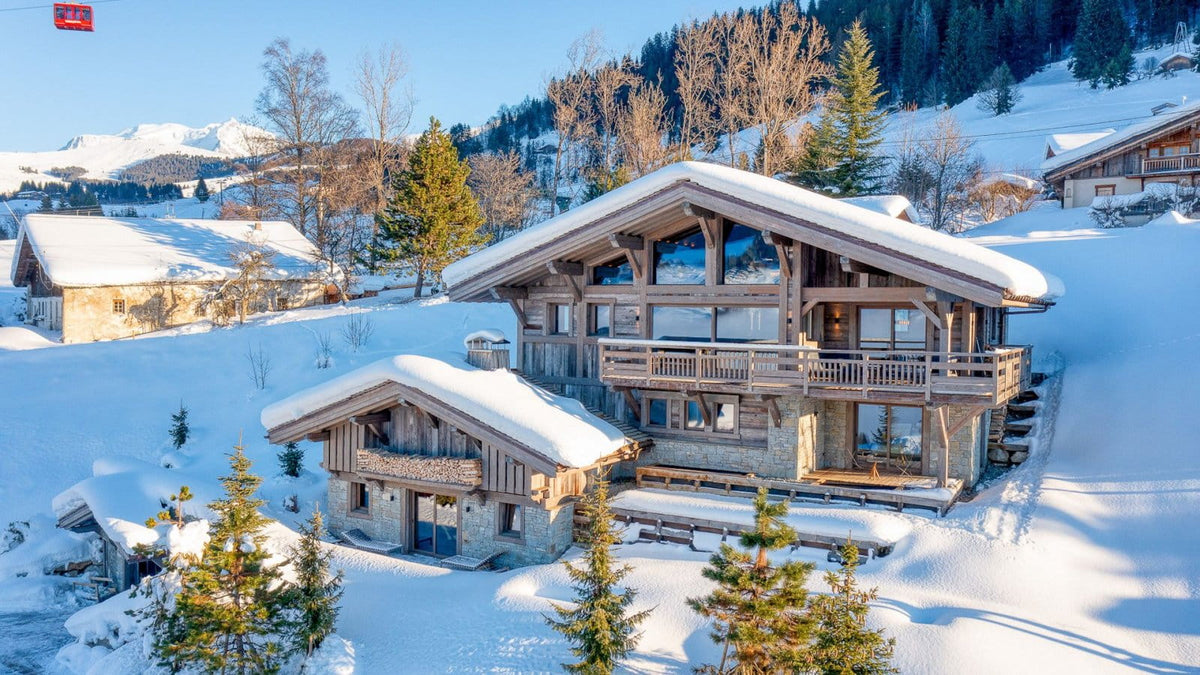 A large alpine chalet with snow-covered roofs and wooden balconies, surrounded by snow-covered mountains, trees, and other chalets under a clear blue sky.