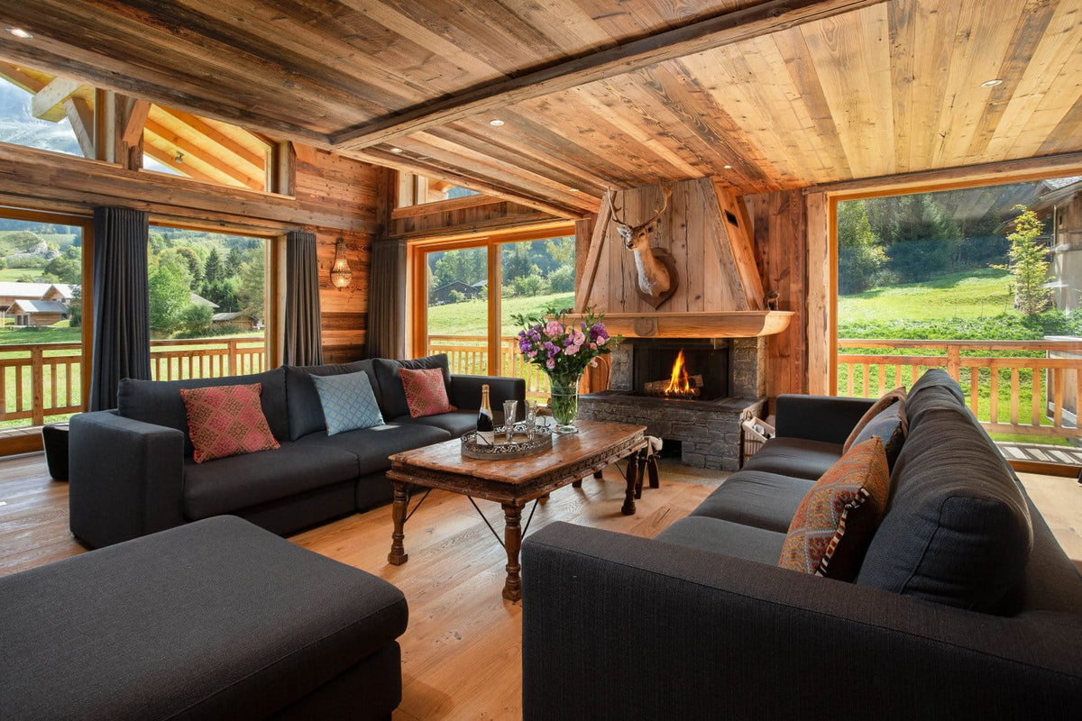 A luxurious mountain chalet living room featuring dark gray sectional sofas, a rustic wooden coffee table, a stone fireplace with a large wood-burning chimney, and large windows looking out onto a green valley.