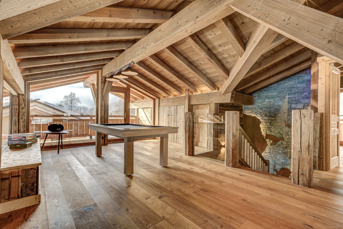 A rustic wooden interior room with exposed timber ceiling beams, wood flooring, a light wood pool table, and a large blue-toned wall mural on the right.