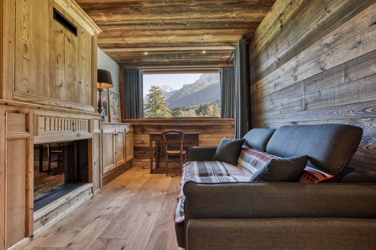 A cozy mountain chalet living room with rustic wood-paneled walls and ceiling, a dark gray fabric sofa with plaid pillows, a built-in fireplace, and a large window overlooking a mountain landscape.
