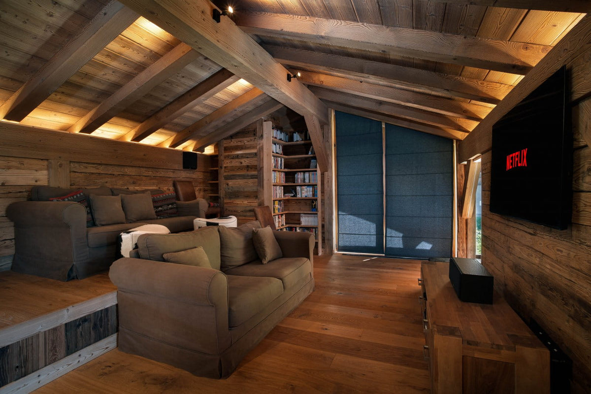 A rustic wooden attic living room with exposed beams, warm ambient lighting, a brown sofa in the foreground, bookshelves, a black TV mounted on the wall, and a Netflix logo visible on the screen.