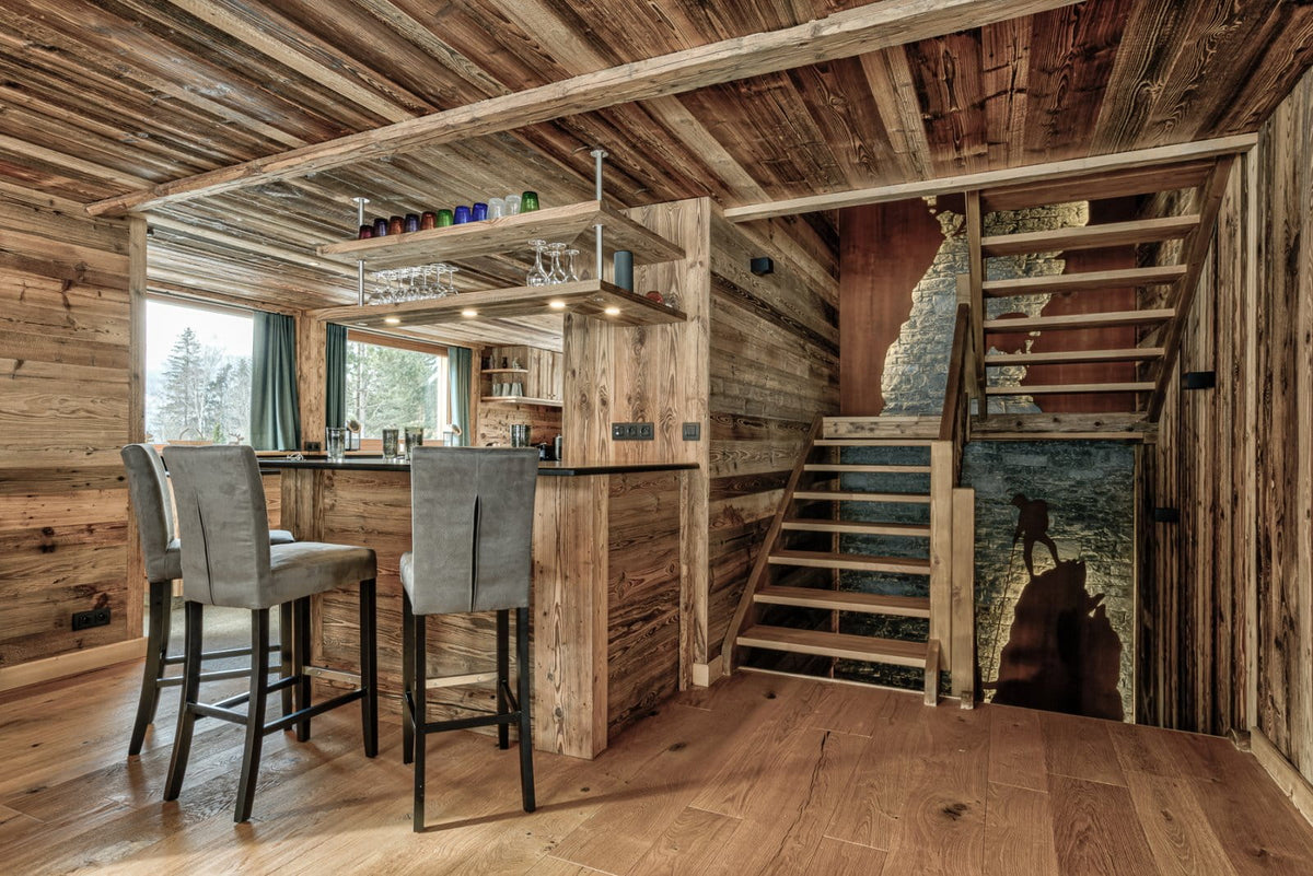 A rustic wooden chalet interior featuring a bar area with three gray upholstered bar stools, a staircase with a stone handrail, and a large wall mural depicting a mountain goat on a snowy slope.