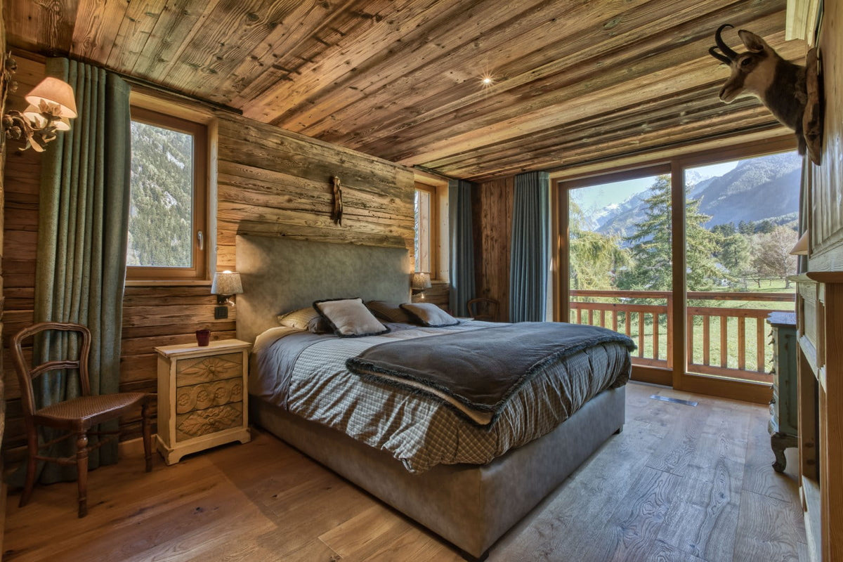 A rustic mountain chalet bedroom featuring a large bed with gray bedding, a wooden nightstand, green curtains, and a view of mountains through large windows.
