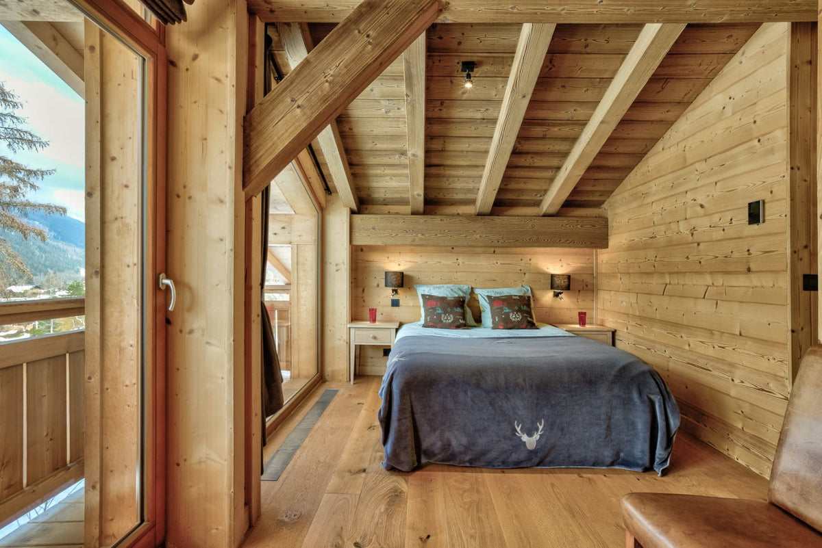 A cozy bedroom interior with a bed dressed in a dark blue blanket and patterned pillows, set in a rustic wooden chalet with exposed beams, wood-paneled walls, and large windows overlooking a mountain view.