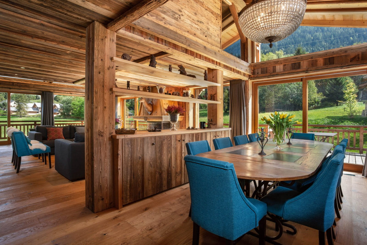 A rustic wooden chalet interior featuring a dining area with a rectangular wooden table and eight teal-blue upholstered chairs, a wooden bar counter with open shelving, and large windows overlooking a green mountain landscape.