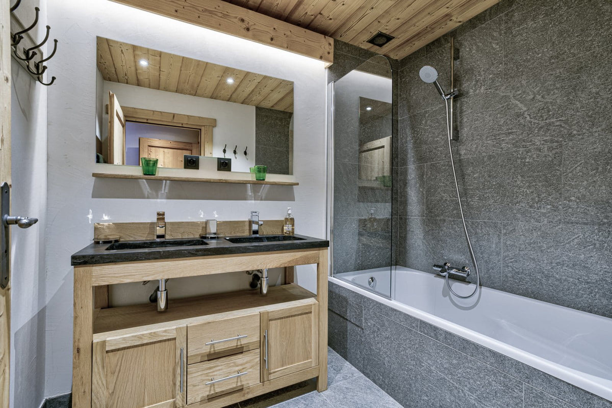 A modern luxury bathroom featuring a double-sink vanity with a black countertop and light oak wood cabinet, a large wall mirror, a white bathtub with a gray stone surround, and a wood-paneled ceiling.