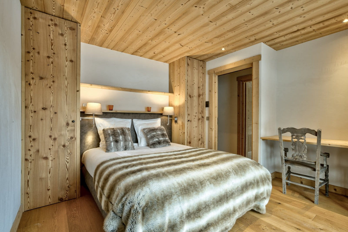 A rustic mountain-style bedroom featuring a large bed with a striped beige and brown faux-fur throw blanket, set against light wood paneling on the walls and ceiling, with a small wooden desk and chair in the corner.