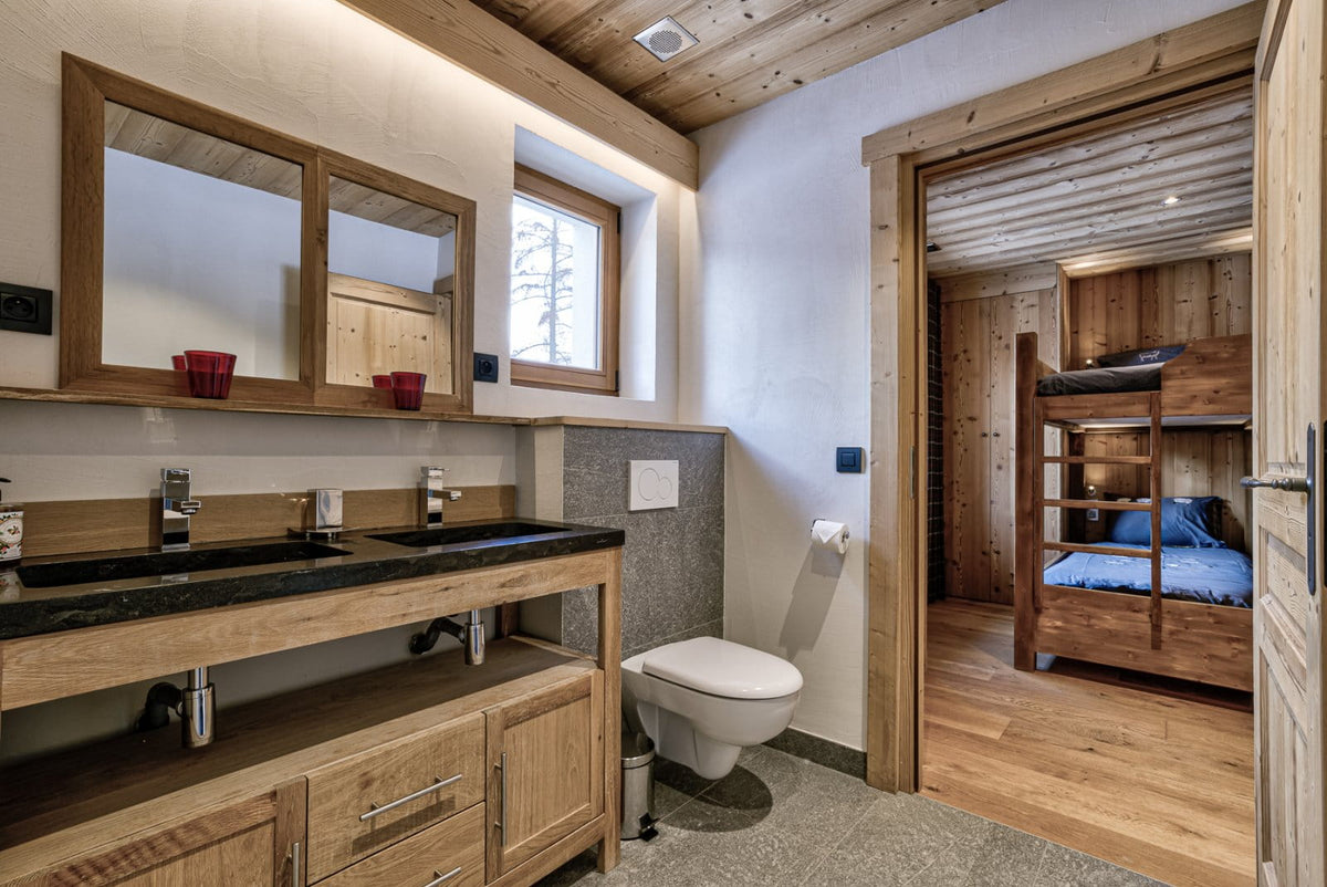 A modern rustic bathroom with a double-sink vanity in light wood, a white toilet, and a large mirror. An open doorway reveals a wooden bunk bed with blue bedding in the adjacent bedroom.